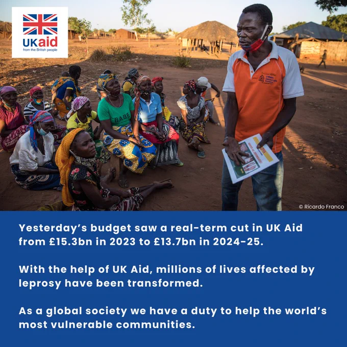 UK Aid worker conducting an educational session with a group of adults in a rural village setting, with traditional huts in the background.