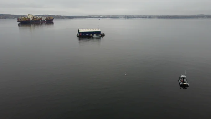 Image d’une étendue d’eau avec un navire de charge en arrière-plan, une structure ressemblant à une habitation flottante au milieu, et un petit bateau en avant-plan. Un petit objet cylindrique est visible au milieu de l’eau.