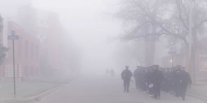 A troop of cadets march down the street. There is fog in the background.