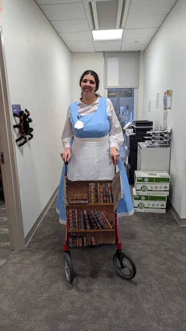 A light skinned Latina woman is wearing a long sleeved white shirt, blue dress on top of it, and a white apron. Her dark hair is pulled back in a low ponytail and in front of her is a walker, which she's holding on to. The walker is covered in cardboard made to look like a library.