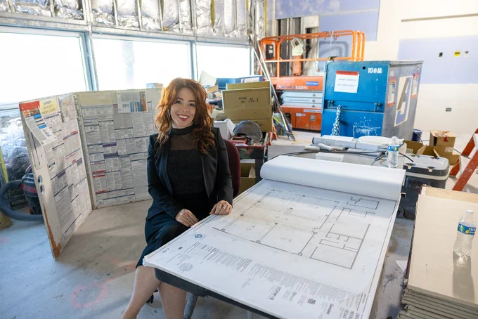 Woman seated at a table in a construction site with blueprints in front of her.