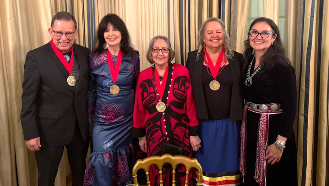 A photograph of five Native and Alaskan Native people wearing traditional outfits. One man and three women wear medals around their necks with red ribbon.