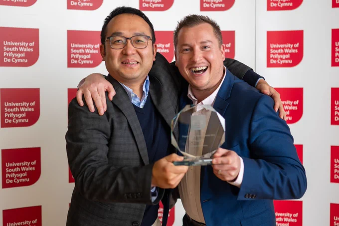 Adam Jones and Kang Li are hugging and smiling and posing with their award. They are both smiling.