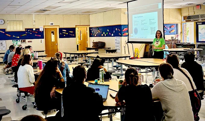 Elementary school cafeteria with tables of teachers facing a projection screen and Mollie in front introducing the session.