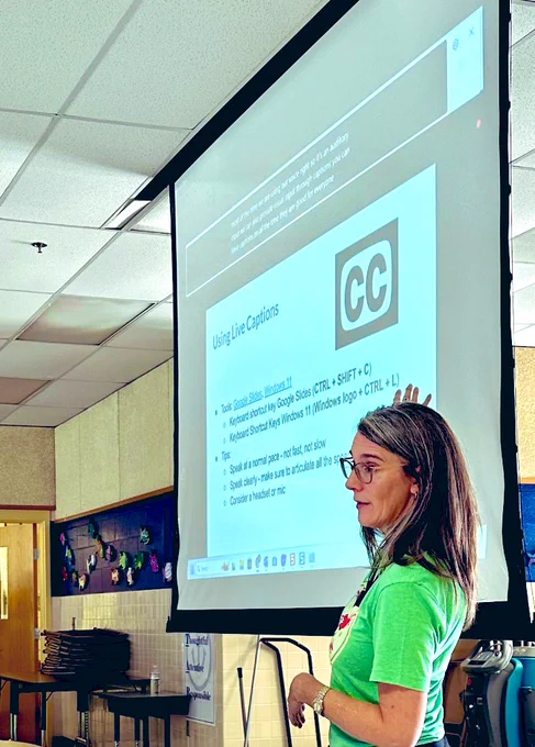 Mollie looking out toward audience and pointing to a large projector screen that has the closed caption icon and options/tips for live captioning.