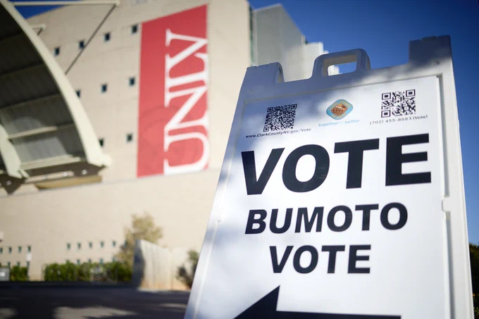 Voting sign with "VOTE BUMOTO VOTE" and a building with a red banner in the background.