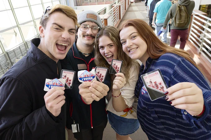 Four young people smiling, holding "I Voted" stickers indoors.