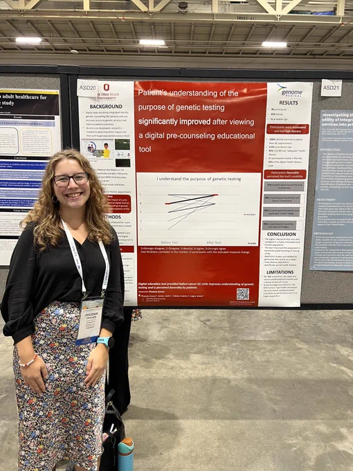 Phoenix is smiling, wearing a black top and a floral skirt. She is next to her research poster.