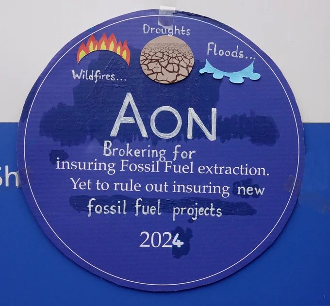 Circular Blue Plaque inscribed with Wildfires, Droughts and Floods. Text says AON Brokering for Fossil Fuel extraction. Yet to rule our insuring new fossil fuel projects 2024. 