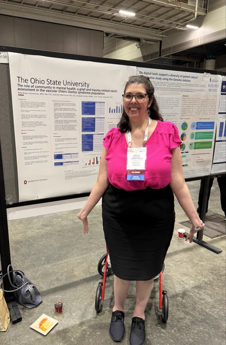 Maya stands to the right of her poster. She is wearing a pink top and black skirt and smiling.