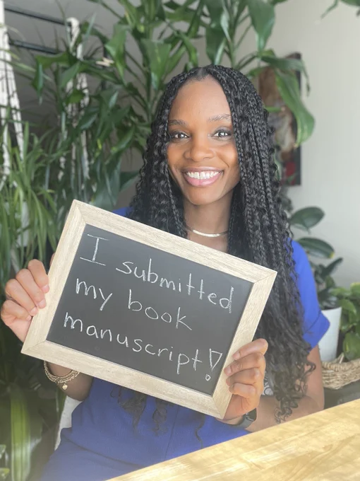 A young Black woman with braids holding up a mini chalkboard that reads, "I submitted my book manuscript!"