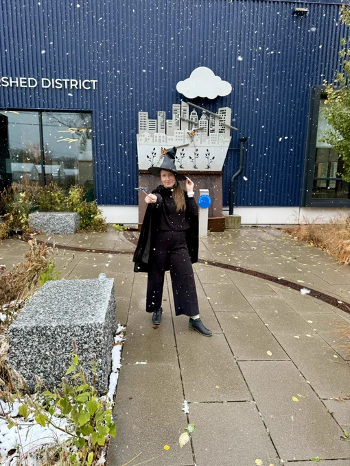 Person wearing black clothing, a black cloak and a black witches hat is posing with a wand pointing at the camera. They are standing outside of a blue clad office building in falling snow. 