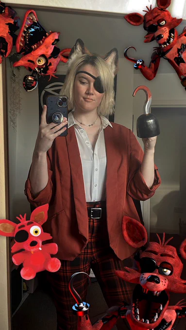 Melissa dressed in a white shirt, red checked trousers, and a red blazer, with an eye patch over her right eye and a silver bloody hook on her right hand. Four photos of Foxy The Pirate from Five Nights at Freddy’s surround her.