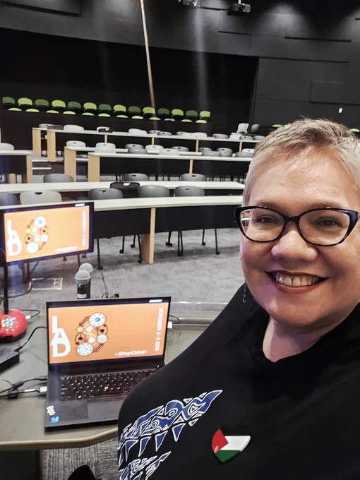 In an empty lecture theatre,  fair-skinned woman, wearing glasses, selfies with the camera. She is wearing a Palestinian Flag pin.