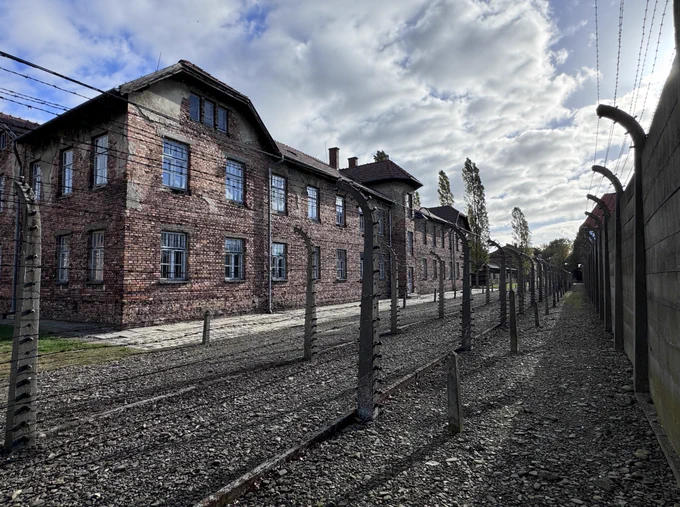 This image displays the exterior of a red-brick building within the former Auschwitz I camp, bordered by rows of barbed wire fencing. There is a wall on the right-hand side. 