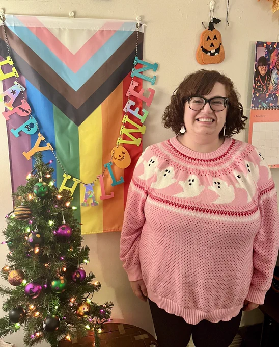 Daniele is a short, fat white woman with short, curly auburn hair and glasses. She smiles in a pink sweater patterned with ghosts, standing with a small tree decorated in Halloween ornaments and a Progress Pride flag. End alt text.