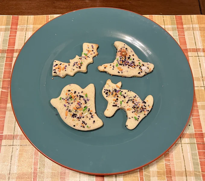 A plate of four sugar cookies in the shapes of a bag, a witch’s hat, a ghost, and a cat. They are decorated with Halloween sprinkles. End alt text.