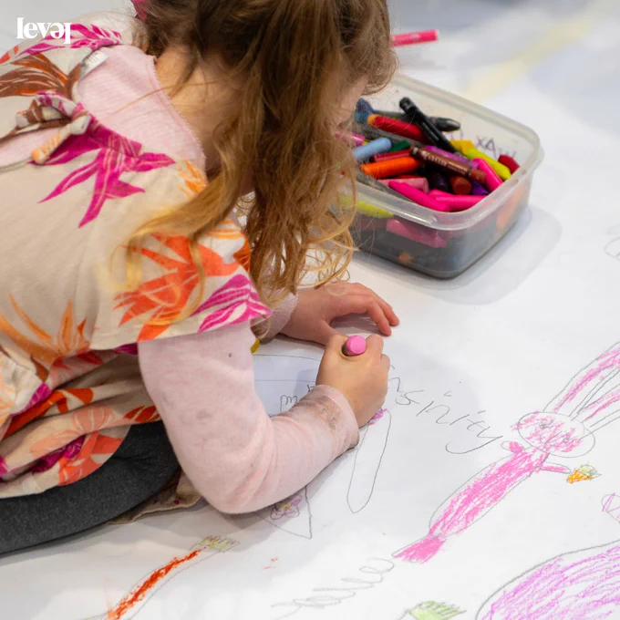 A young person kneels down on a sheet of white paper. They are drawing and colouring with crayons. They have drawn a cute pink bunny rabbit. The wear a pink and orange flowery dress and have long brown hair in bunches.