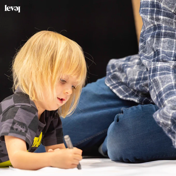 A young person sits on the floor drawing onto a large sheet of white paper. An adult sits behind them but you can't really see the adult. The young  person has shoulder length blonde hair.