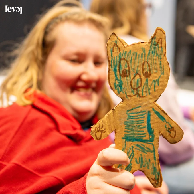 A person smiles and holds up a cat that they have cut out of cardboard. They have coloured the cat in with blue felt tip pen. The person is out of focus in the photo. They wear a red top and have shoulder length blonde hair pulled back from their face with a headband.