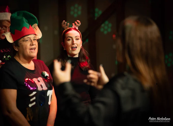 Two women sing with joy in festive attire, one wearing an elf hat, the other reindeer antlers, under warm holiday lights.