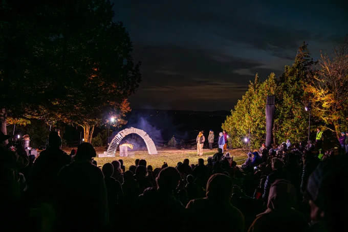 Night has fallen and the arch stands completed. A fire is burning in an oil drum directly underneath it, as singers Aminita, Phil, Ellian and Kate stand vigil to one side of the arch. Hundreds of audience members sit and stand all around it, watching the ice melt.