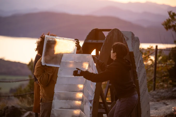 As the two builders near the top of the arch, they stack the last ice blocks in place. Nao lifts one with both hands, while Irene braces herself to receive it. 