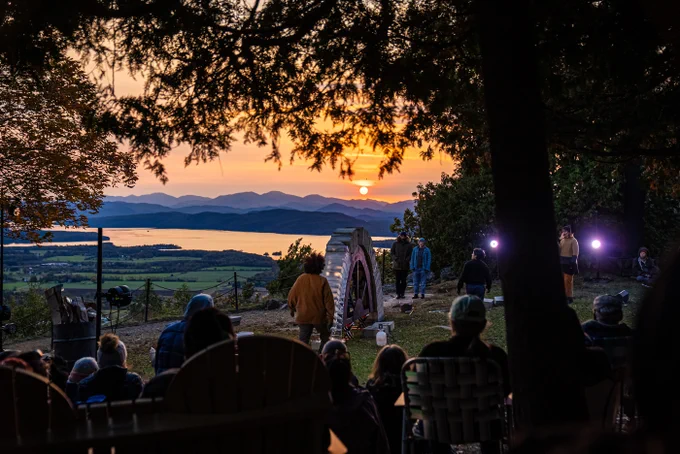 The summit of Mount Philo in Vermont, USA at sunset. A crowd of people, silhouetted against the light, watch as two performers walk towards an arch. It is about 2 metres tall and is partially built, made of two thirds concrete blocks and one third ice, supported by a wooden former. 