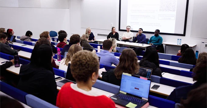 Attendees of a University College London Careers Service event, sat in a lecture theatre. There are panellists sat in front of them, behind a table. Behind the panellists is an electronic screen, displaying a slide show.