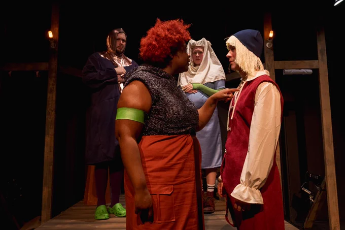 A scene on stage from Beards. Four people in medival dress stand on a darkened stage. At the forefront of the photo, a person points at the chest of another character while the other two people look on. 