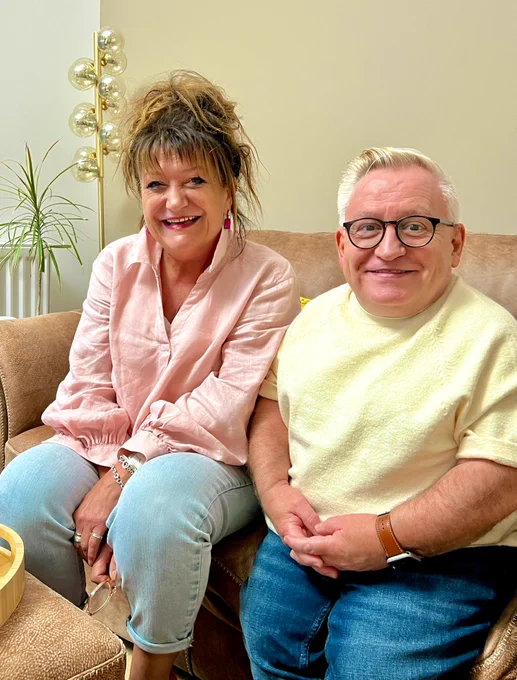 Jane on the left, pink blouse, pale blue jeans, Simon on the right, yellow top and mid blue jeans. Both sat on a brown leather sofa with a plant and light with many bulbs behind them. 