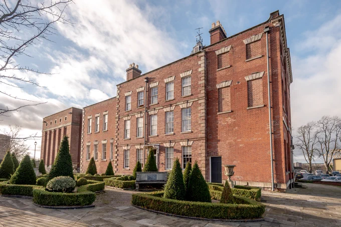 A large red-brick building with trimmed bushes in front of it. The weather is sunny with some clouds.