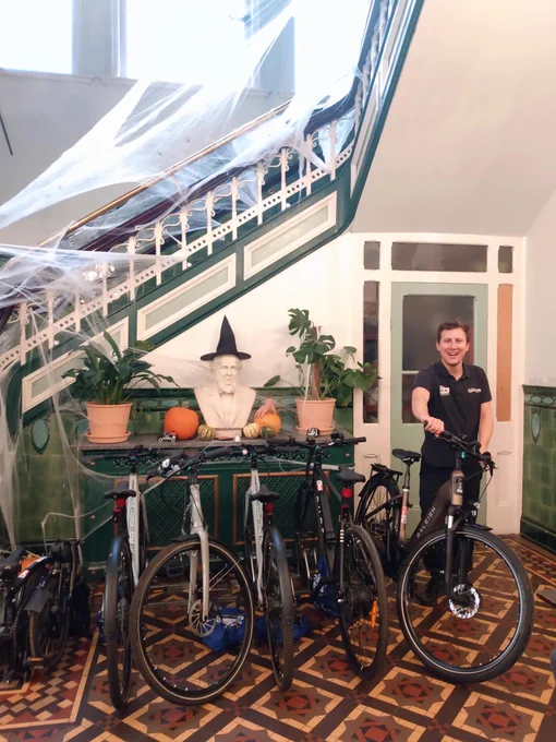 Cycling UK staff member Will is standing next to a rack of five e-bikes and one electric Brompton, on a beautiful brown, cream and red tiled floor.  Behind him is a marble bust wearing a Halloween witch's hat and surrounded by pumpkins.  Behind that is an ornate staircase decorated with cobwebs for Halloween.