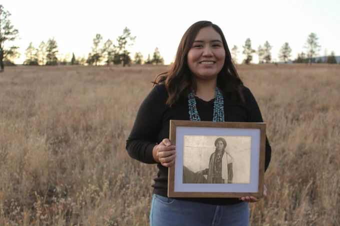 A young Native woman with a turquoise necklace holds a black and white photo of a young woman.  