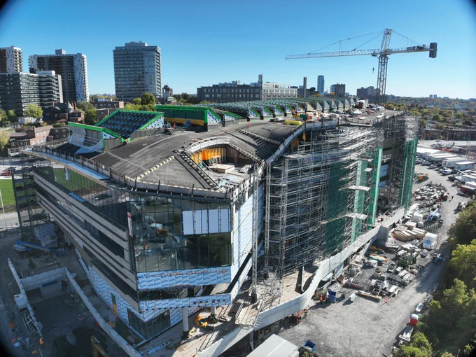 Photo prise par un drone de l’installation Ādisōke; on y voit les lignes incurvées de la toiture en bois et les fenêtres finies. À l’arrière-plan, on aperçoit des immeubles de grande hauteur et une grue de chantier.
