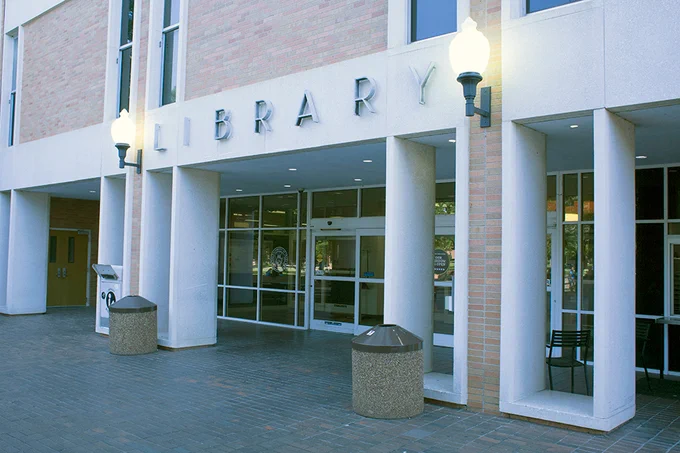 Entrance of a library building with large glass doors, flanked by outdoor lamps and trash receptacles.