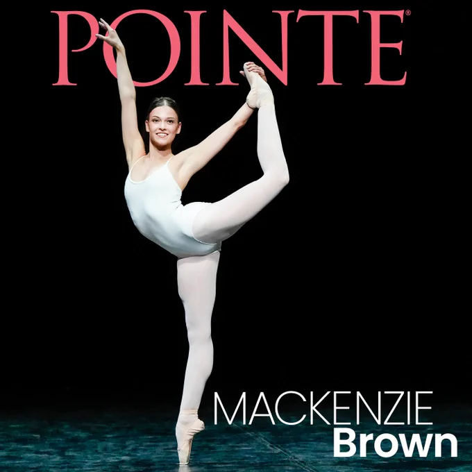 Image description: Mackenzie Brown grins warmly as she stands on point while holding her left foot in a high attitude. She wears a white unitard and a sleek high bun with a middle part. Behind her is the Pointe logo in bright pink, in all caps, and her name in white block letters.