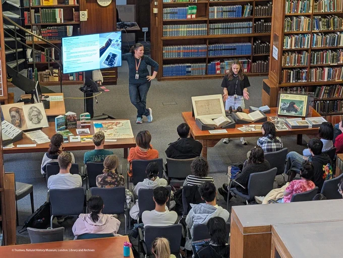 A group of students seated in a library reading room, watching a presentation by two staff. There are two large desks with a display of items on.