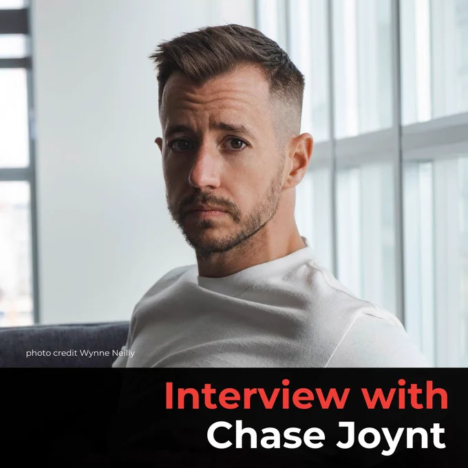 A photo of Chase Joynt, a light-skinned man with short cropped hair and facial hair. He is wearing a white t-shirt and looking into the camera.