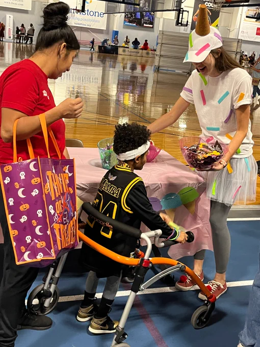 A woman dressed in an ice cream cone costume handing out candy to a little boy who is using a walker and dressed as a basketball player. His parent stands behind him holding  a trick or treat bag