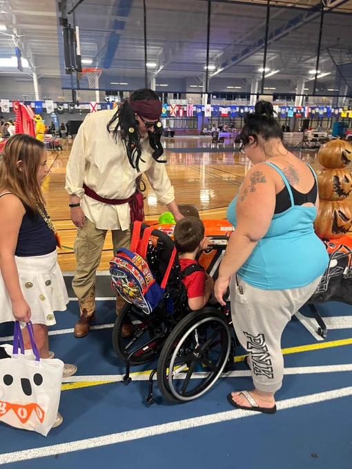 Man dressed as a pirate handing out candy to a boy in a wheelchair. Another little girl stands in line behind him and his parent is next to him. 