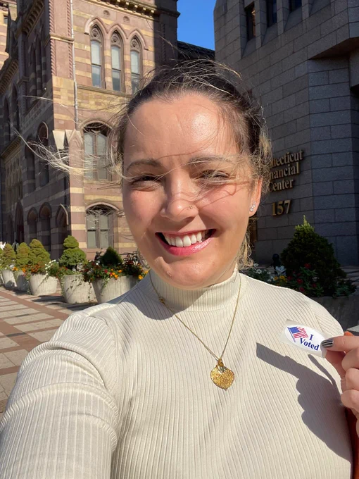 Haley takes a selfie outside of the New Haven city hall. She is smiling at the camera holding an "I voted" sticker. She is wearing a white turtleneck shirt, has a gold aspen leaf necklace, and her brown hair is pulled up. 