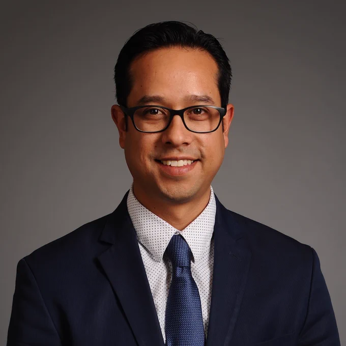 A headshot photo of Aaron Cortes wearing dark colored suit with blue tie and collard shirt and glasses, smiling, against a gray background.
