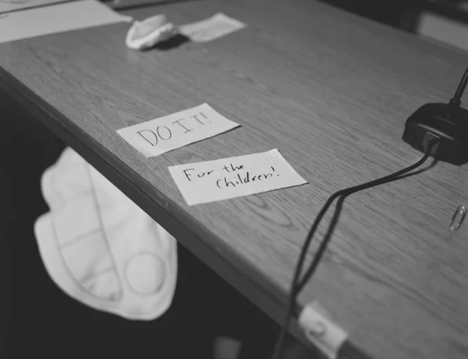 A greyscale photo from Desert Bus for Hope 2023, of the comms table. It is empty except for two papers reading "DO IT" and "FOR THE CHILDREN." A stuffed doll is under the table with a large smile.