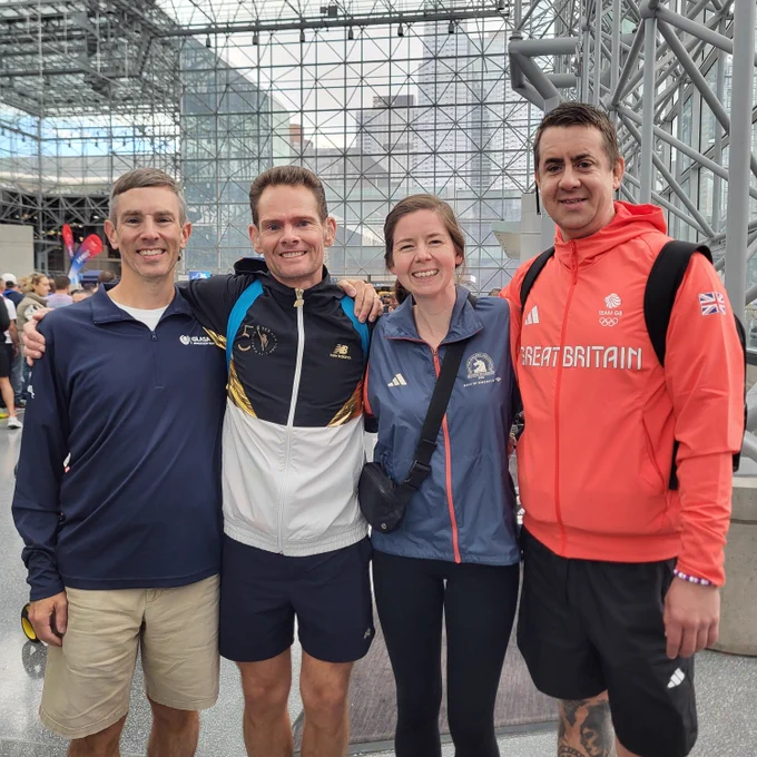 Three white men and a white woman smiling stand arm-in-arm wearing various running jackets. In the background is glass and metal structure that is the interior of the Jacob Javits Convention Center.