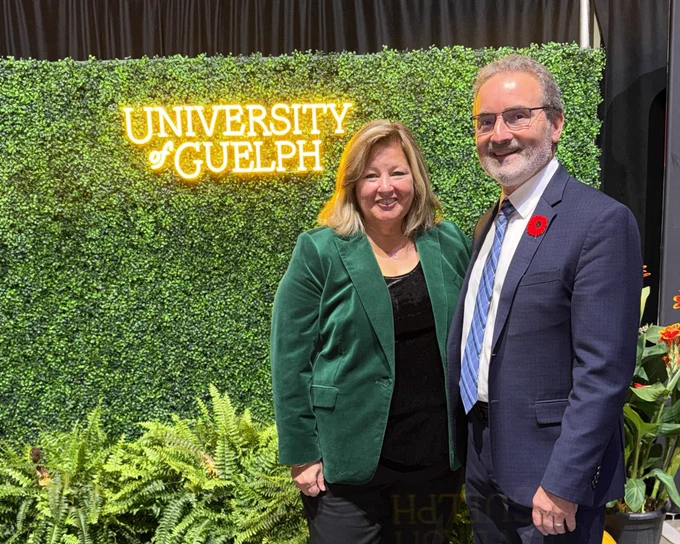 Ontario Minister of Rural Affairs Lisa Thompson and University of Guelph Acting President, Rene Van Acker, smile in front of a wall of fake green plants with a yellow neon sign that reads “University of Guelph”.