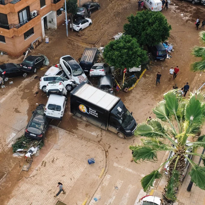 Aerial view of a flooded urban area with several cars submerged in muddy water. People are visible assessing the situation. A recognizable truck from World Central Kitchen is parked among the cars in Paiporta Spain near Valencia