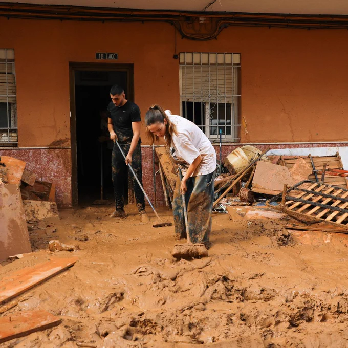 Two individuals are cleaning up mud and debris in front of a building with a noticeable quantity of silt covering the ground and piled belongings in Paiporta Spain near Valencia