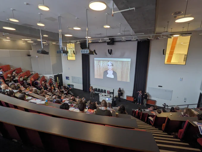 A photo of a lecture theatre full of people watching a woman on a screen deliver a video message
