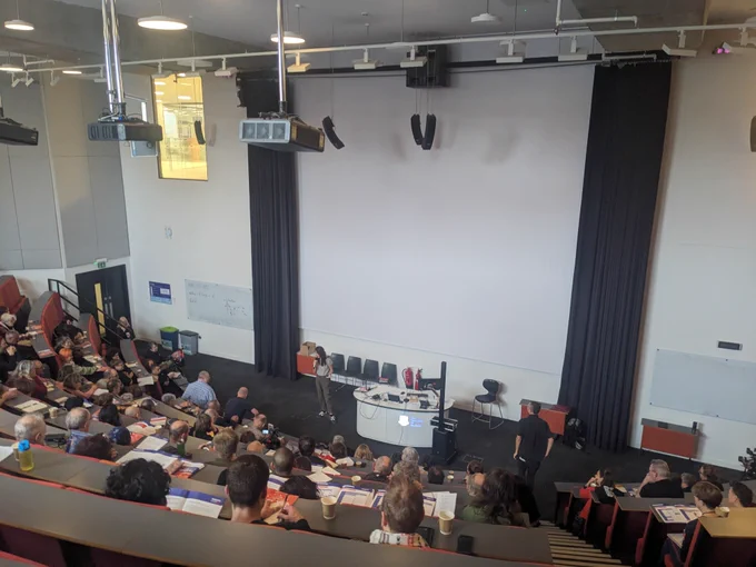 A full lecture theatre of people watching a speaker with a microphone at the front talking